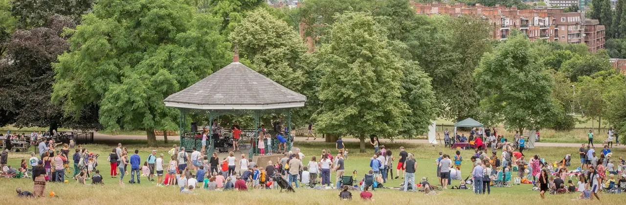 Large crowd of visitors and families gathered on a grassy park with a bandstand, surrounded by trees and Georgian buildings in the background.