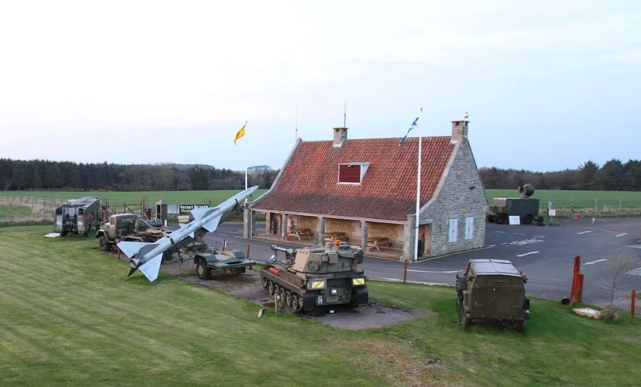 Stone cottage with red roof surrounded by military vehicles and equipment in rural countryside setting.