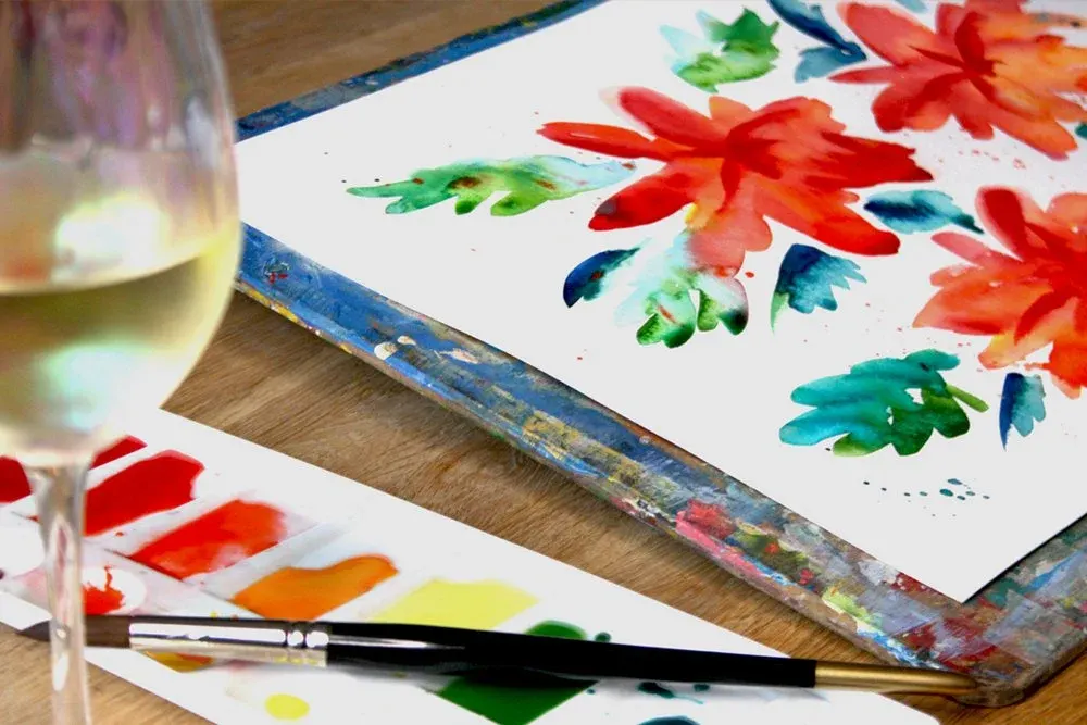 Watercolour paintings of red and colourful flowers on paper with paintbrush, palette and glass of water on wooden table.