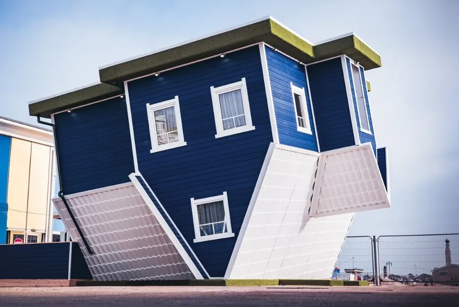 Upside-down blue and white house structure with windows, an unusual architectural attraction at a seaside location.