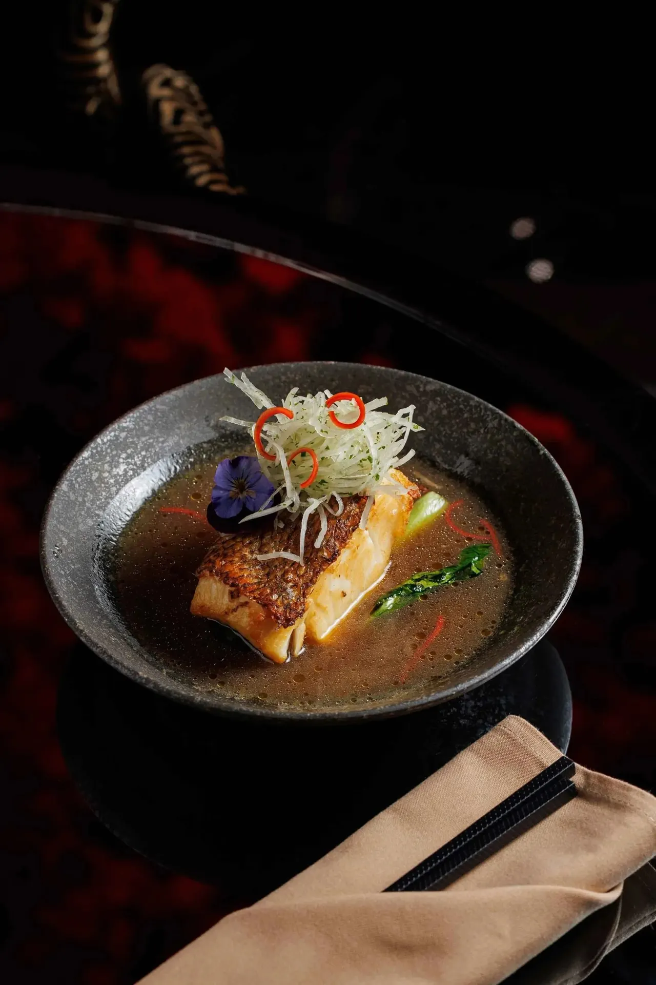 Seared fish fillet in rich broth, topped with julienned vegetables, red chilli rings and an edible flower, served in a dark bowl.