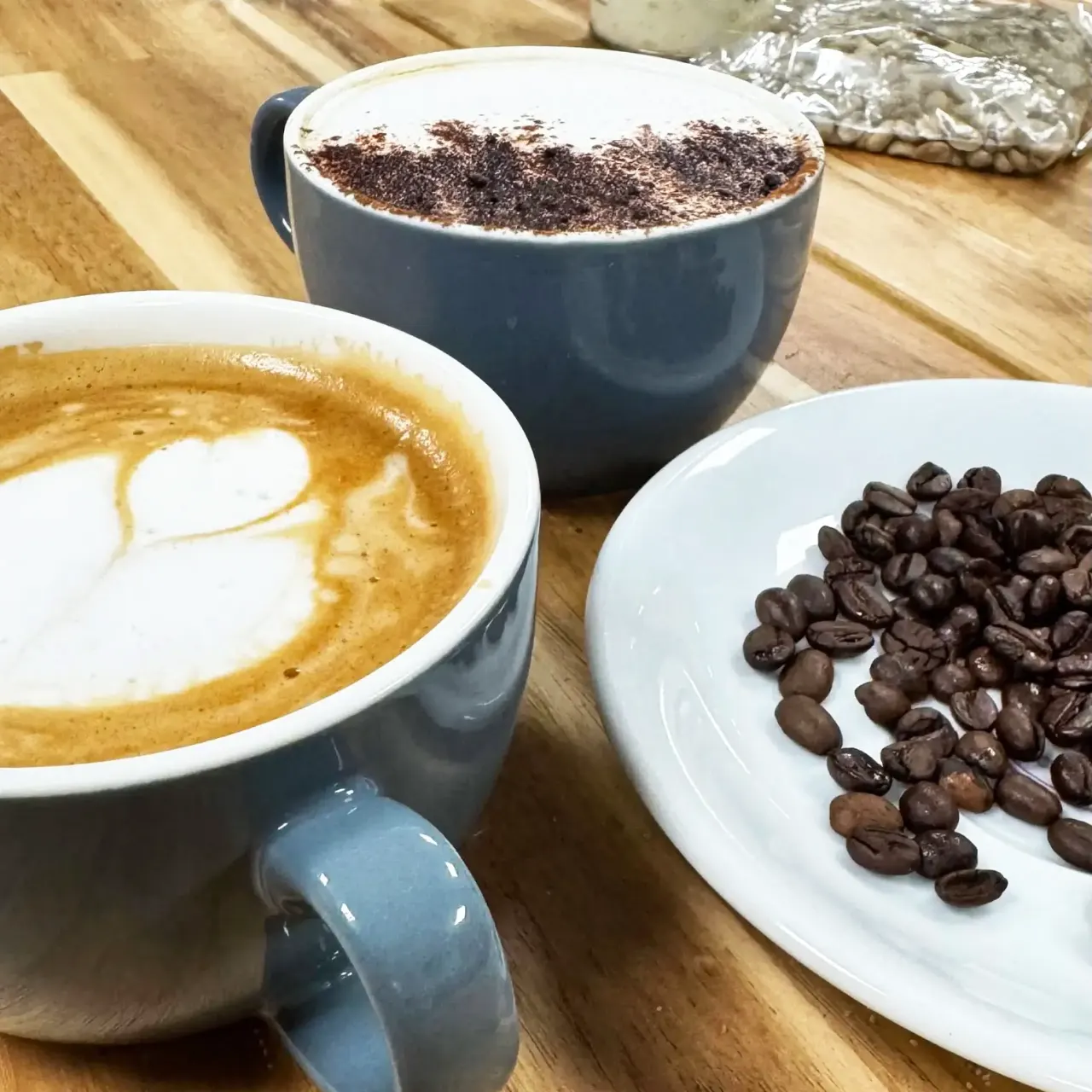 Three coffee drinks displayed on a wooden counter: a latte with latte art, a cappuccino dusted with cocoa powder, and a cup of coffee beans beside them.