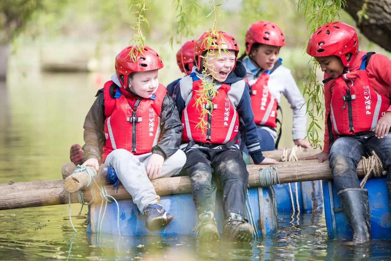 Children wearing red helmets and life jackets sitting on a log raft in shallow water during an outdoor activity.