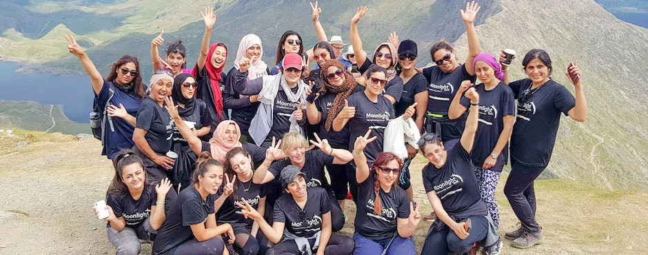Large group of women in matching black t-shirts posing cheerfully on a mountainside with a lake and peaks visible behind them.