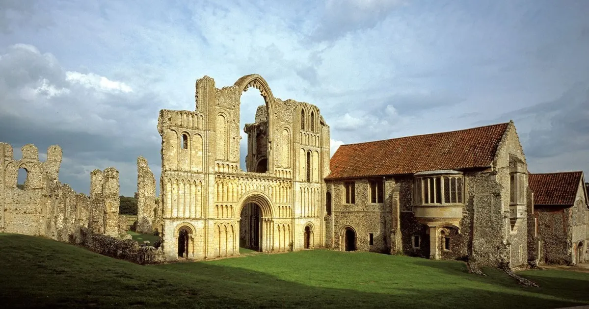 Ruined medieval priory with stone walls and arches set in green grounds under cloudy sky.