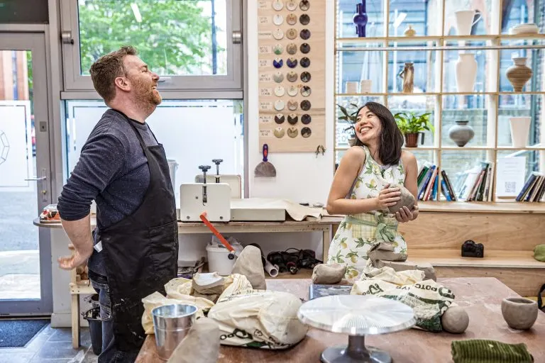 Two people demonstrating pottery techniques at a workbench surrounded by ceramic vessels and tools in a bright studio space.