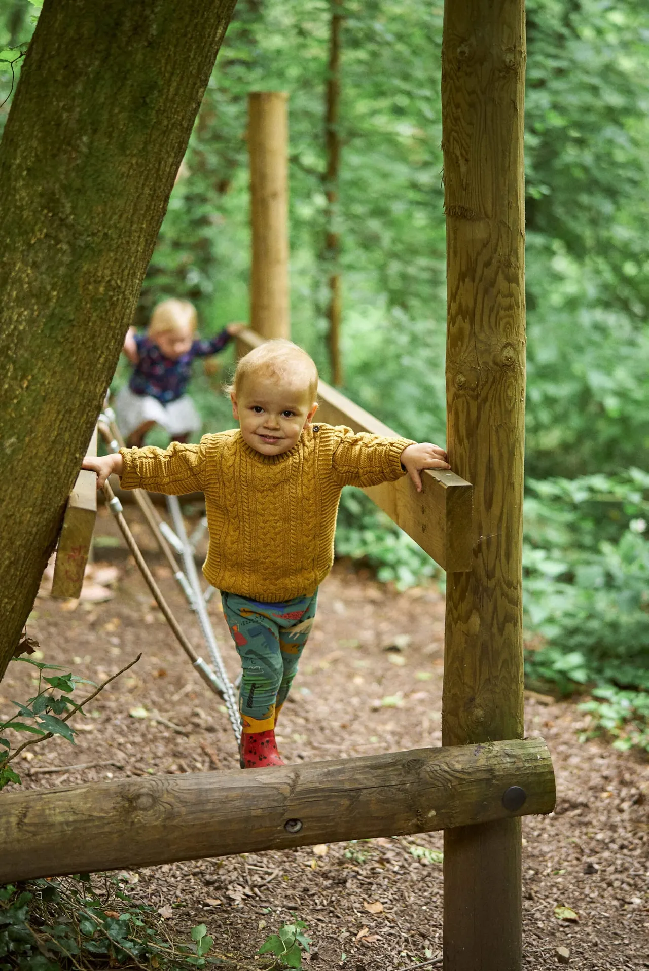 Toddler in yellow jumper balancing on wooden beam in forest playground with another child visible behind.