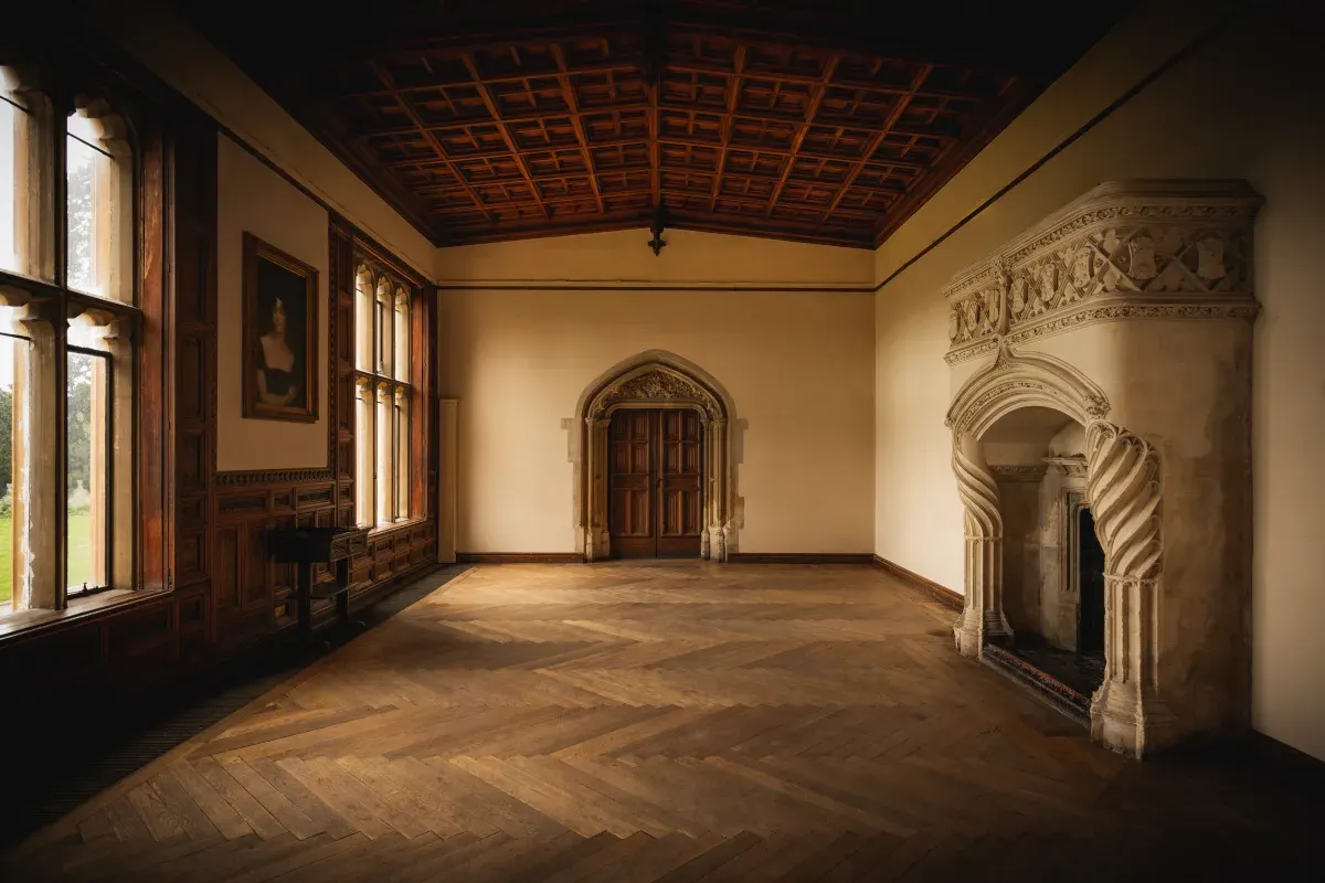 A grand empty room with ornate plasterwork, wooden herringbone flooring, coffered ceiling and tall windows with wooden frames.