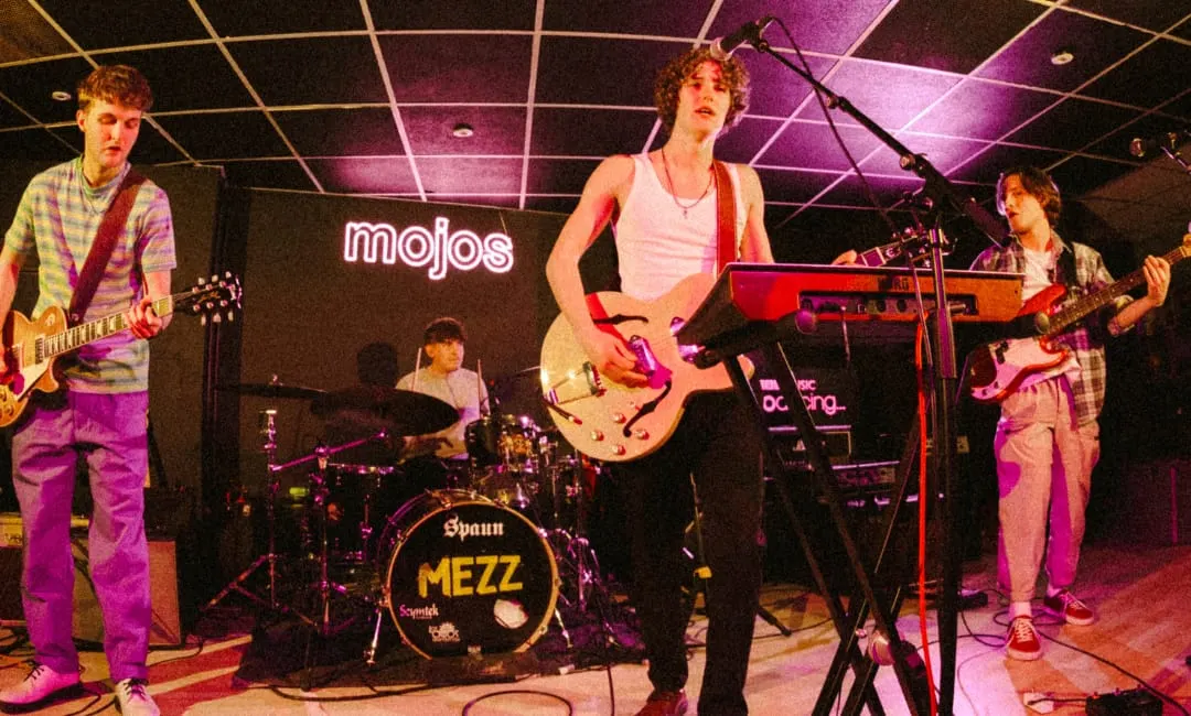 A rock band performs on stage under pink neon lighting at an indoor music venue.