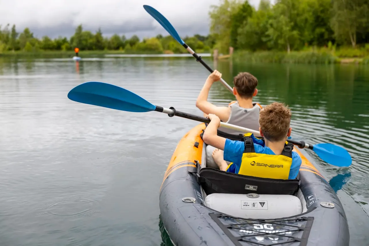 Two children paddling a yellow and black inflatable kayak on a calm river, wearing life jackets.