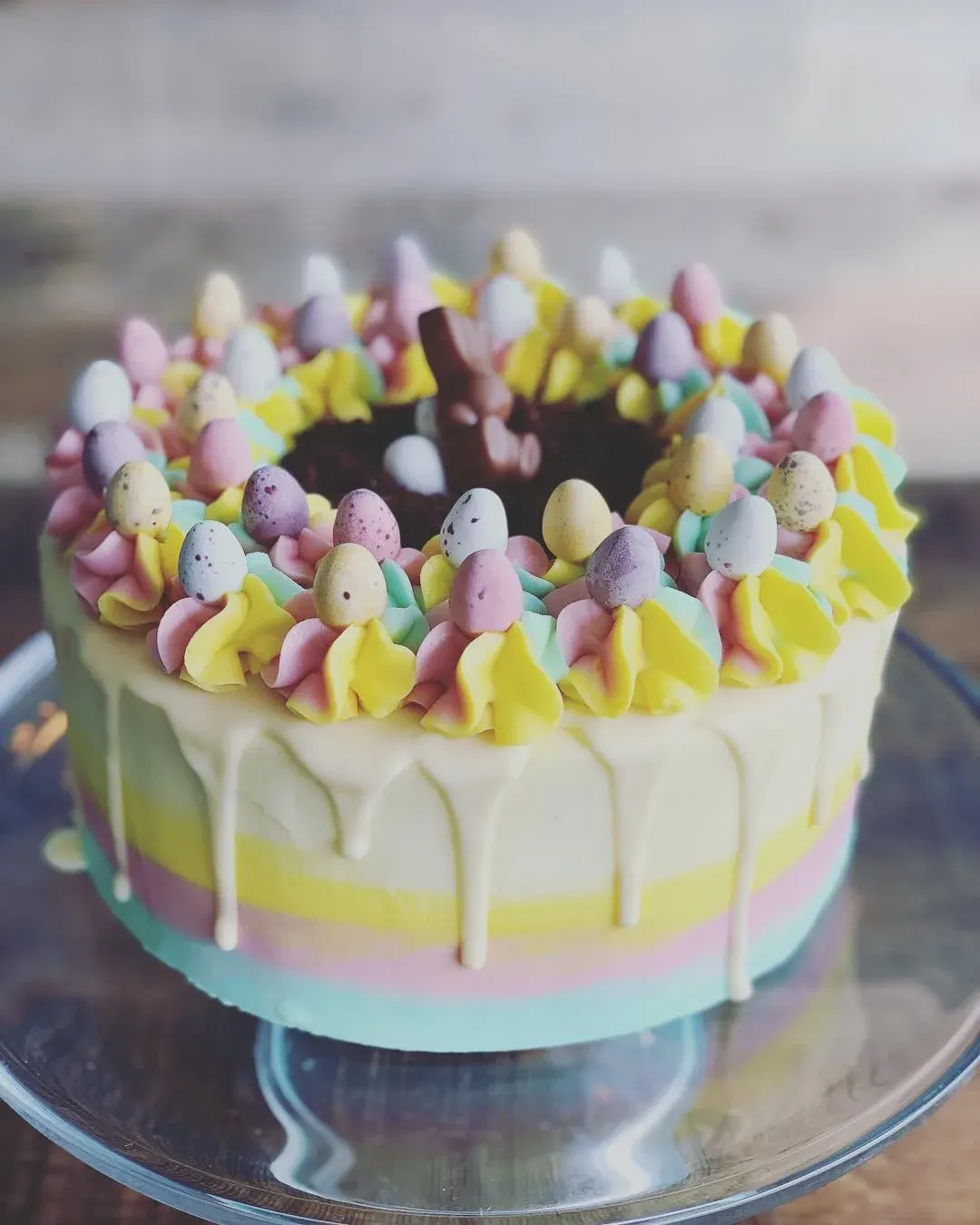 Easter-themed cake with pastel-coloured layers, white drip frosting, and decorative piped rosettes topped with mini speckled eggs.