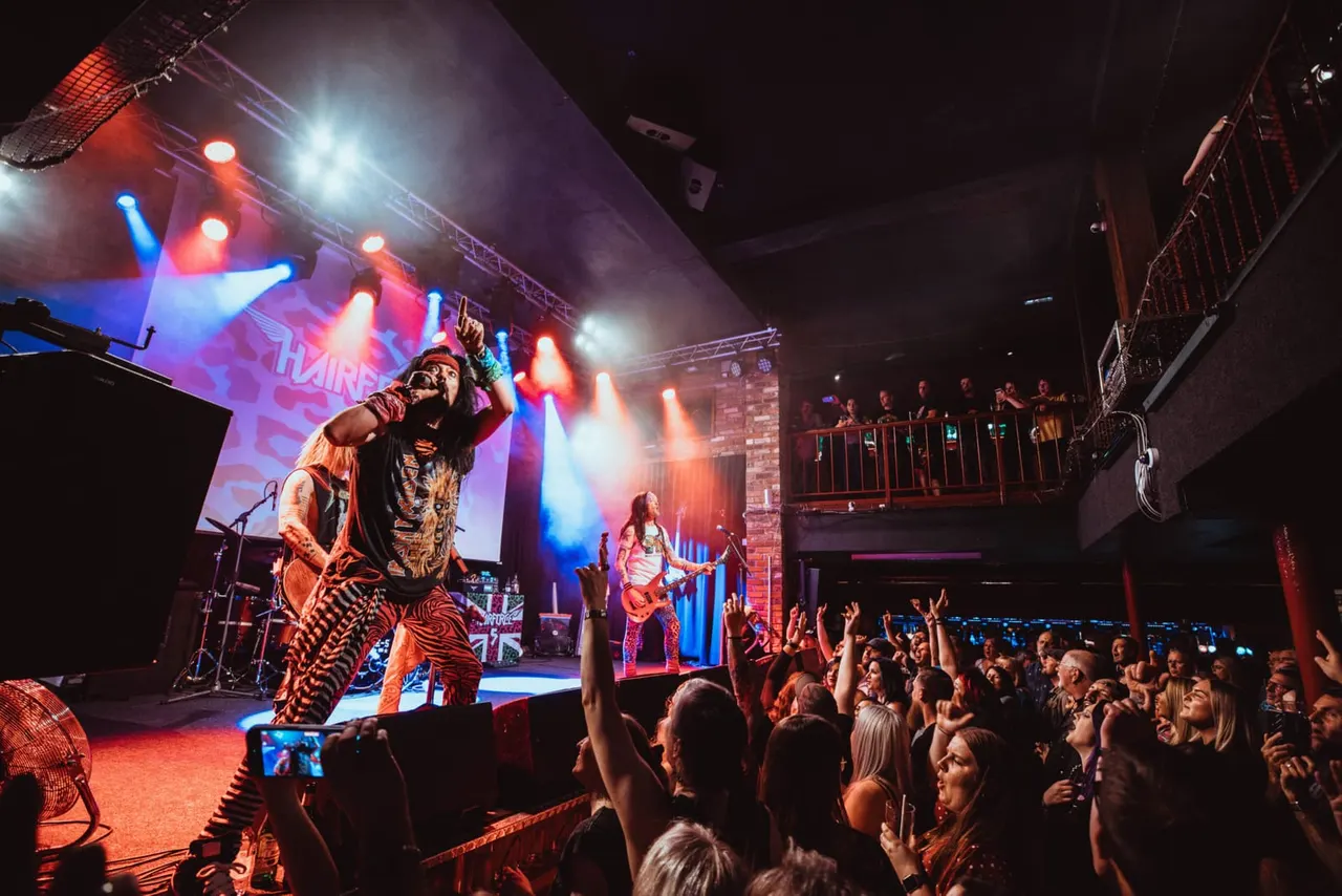 A live rock band performs on stage with colourful lighting whilst a large crowd of audience members watch and hold up phones to record.
