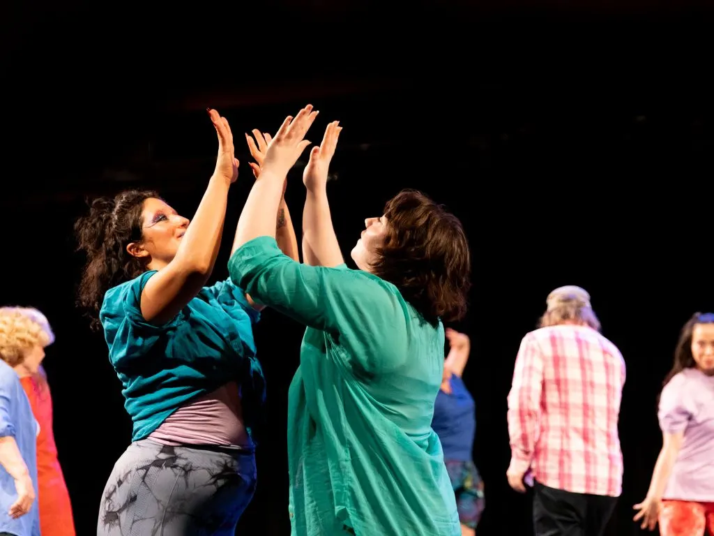 Two women dance together on stage with arms raised, whilst other participants watch from the sides.