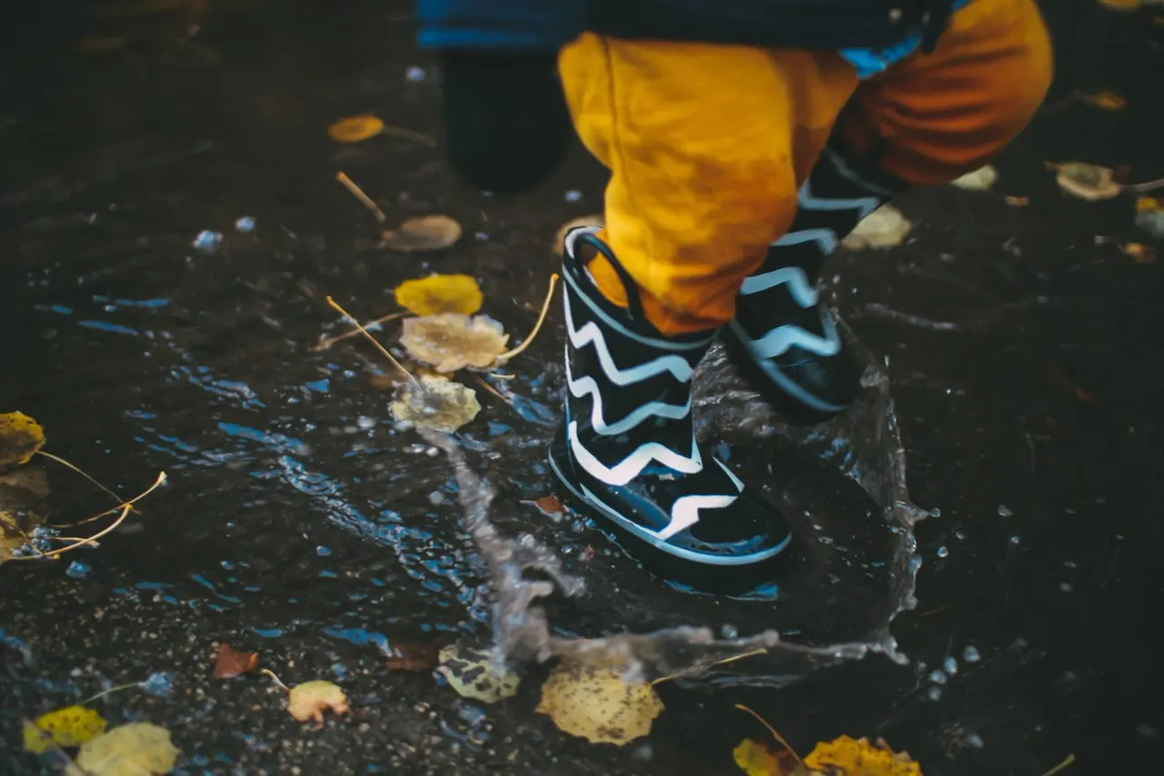 Child wearing colourful patterned boots splashing in a shallow puddle with fallen leaves.