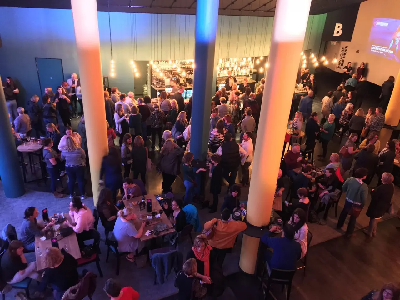 Overhead view of a busy indoor event space with crowds of people socialising, illuminated pillars, and a bar area in the background.