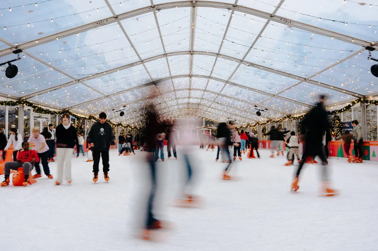 Visitors ice skating indoors under a glass roof decorated with fairy lights and festive garland.