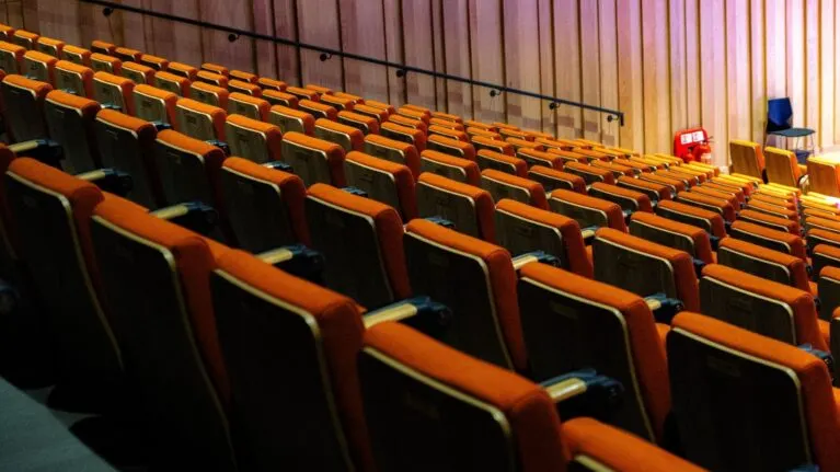 Rows of empty red and orange cinema seats in an auditorium with a exit sign visible.