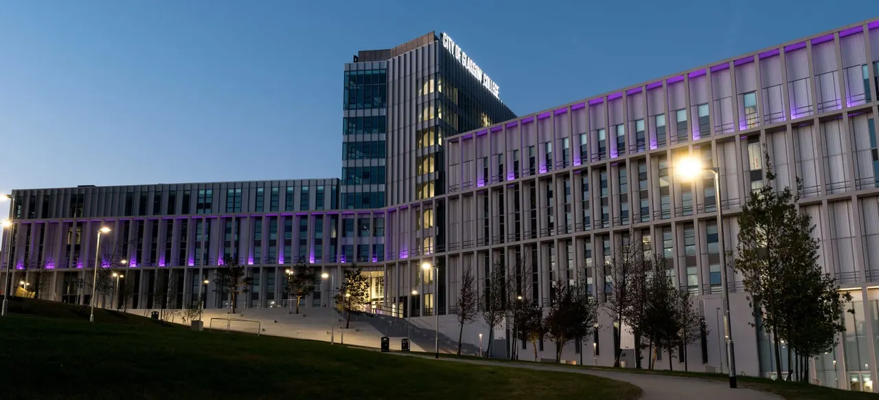 Modern multi-storey building complex with purple illuminated features at dusk, surrounded by landscaped grounds.