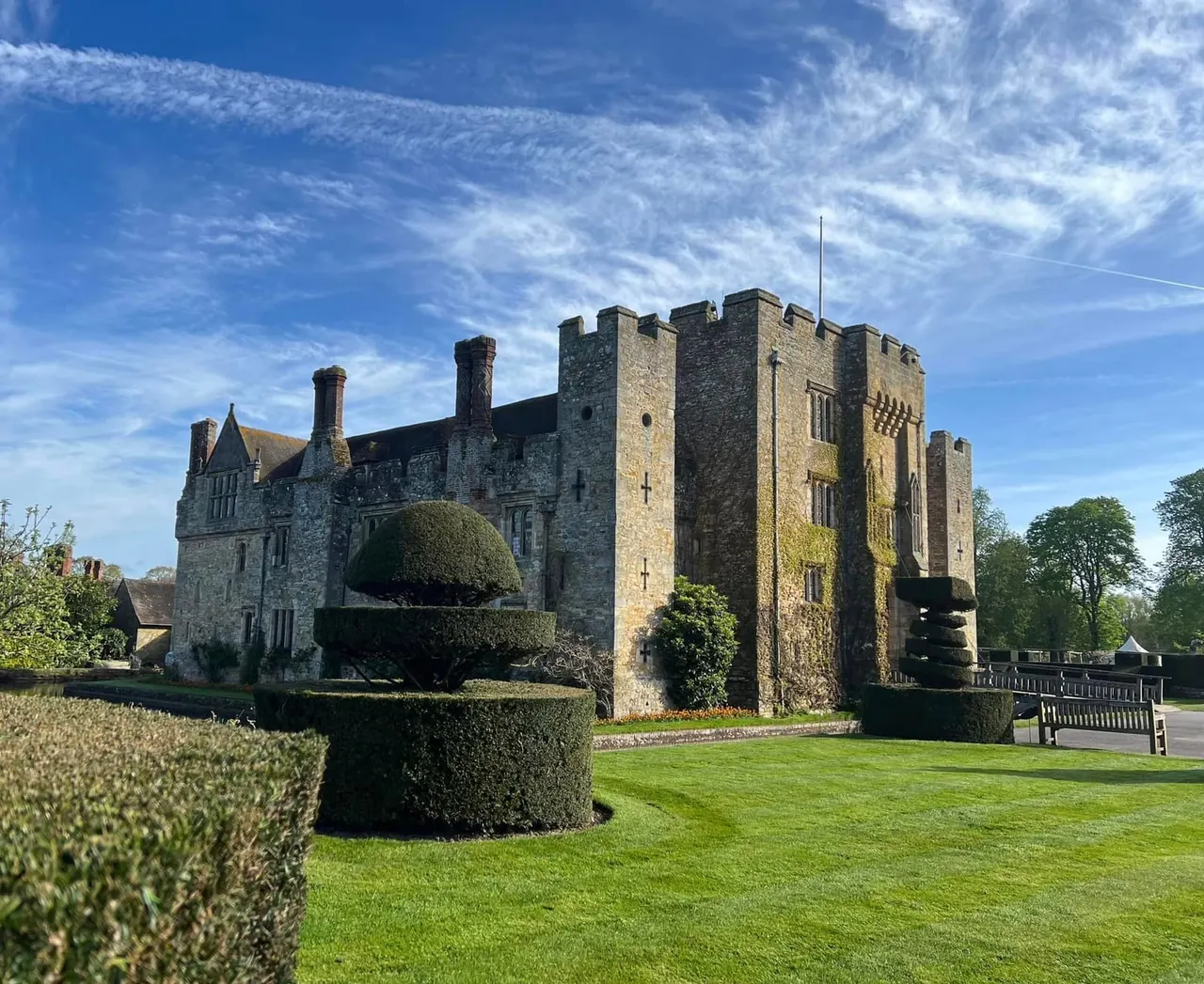 Medieval castle with crenellated towers set in manicured gardens with topiary hedges and green lawn under blue sky.