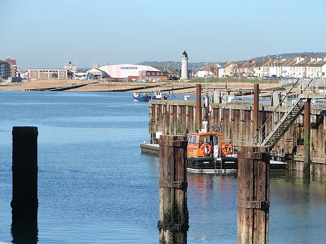 Orange and black workboat moored at wooden pier with coastal town and lighthouse visible across the bay.