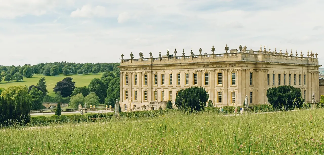 A grand Palladian stately home with ornamental grounds viewed across a wildflower meadow, with woodlands beyond.