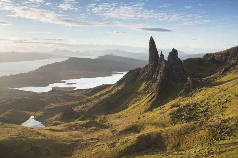 Rugged Highland landscape with a distinctive rock pinnacle, green moorland, lochs and distant mountains under cloudy sky.