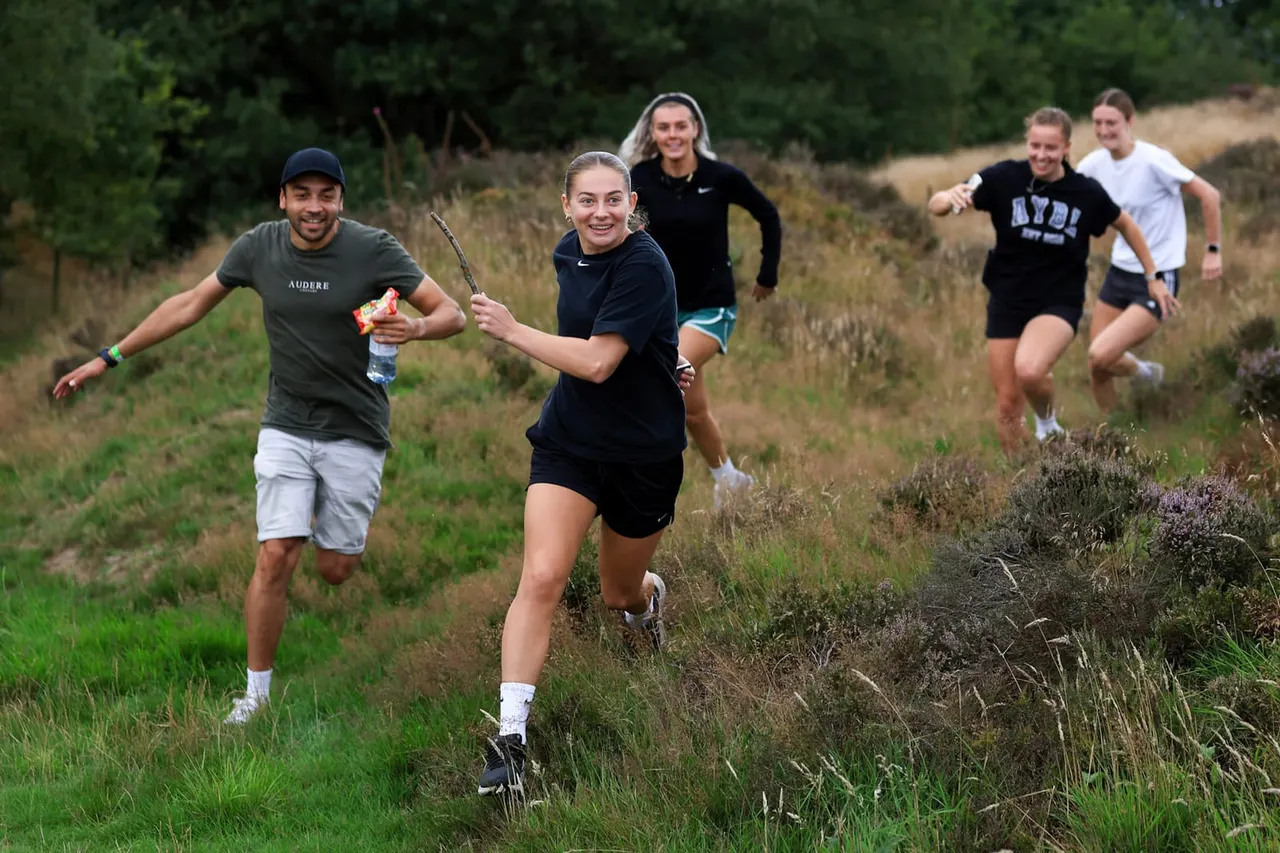 A group of six people running through a grassy, heathland terrain with shrubland in the background during an organised outdoor activity.