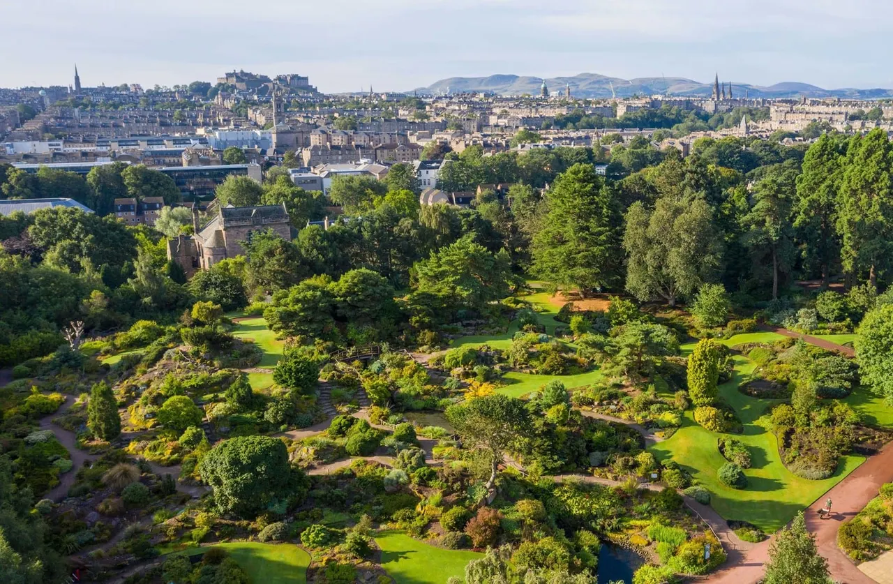 Aerial view of a botanical garden with mature trees, landscaped grounds and a small stream, with Edinburgh's cityscape and hills beyond.