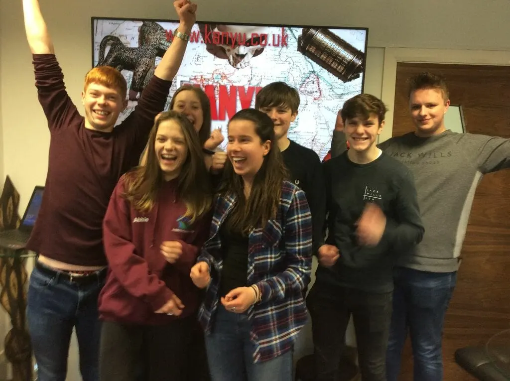 Group of young people celebrating inside an escape room, posing in front of a wall display with a map and trophy.