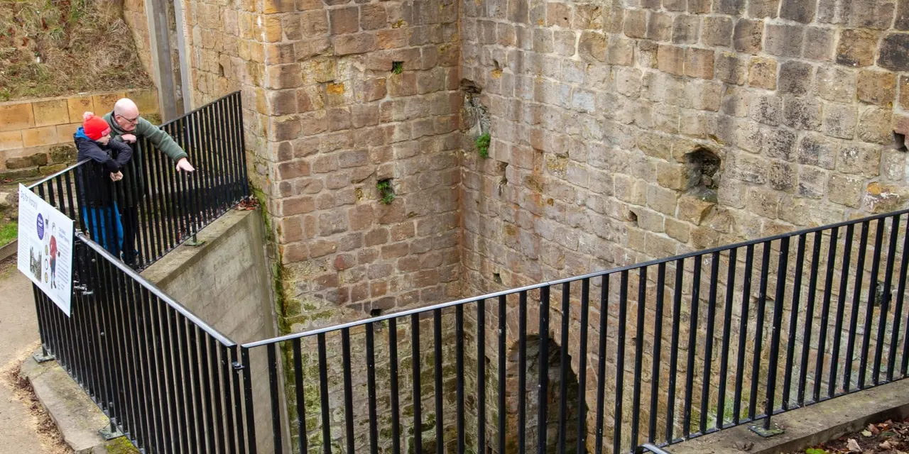 An adult and child in a red hat walking on a metal platform within the stone ruins of a castle, with moss-covered brick walls surrounding them.