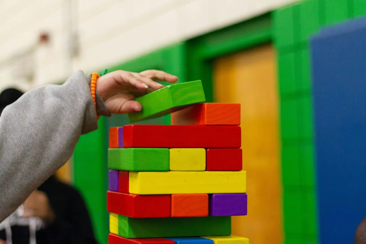 Child's hand placing a green wooden block on a stacked tower of colourful building blocks.