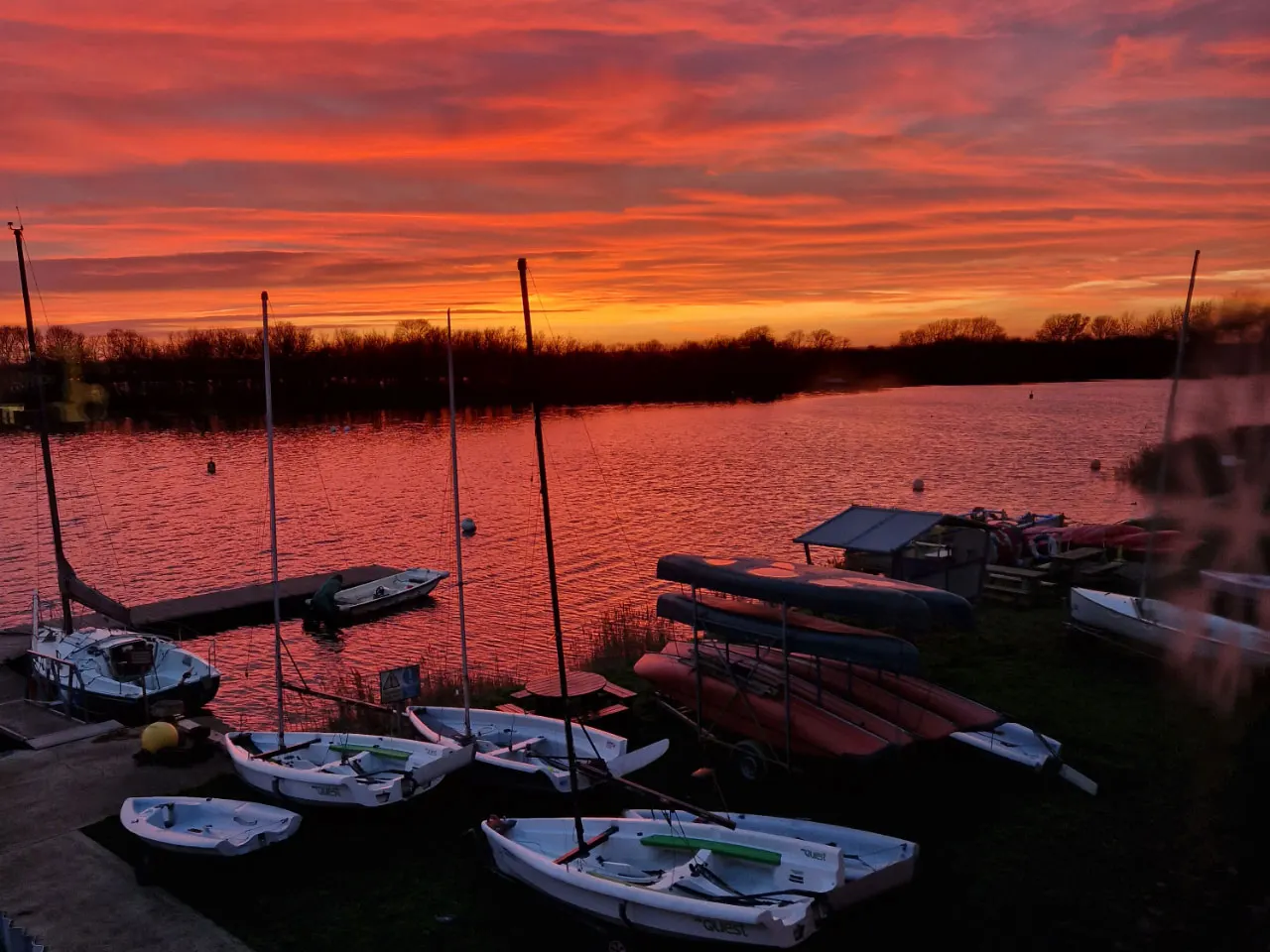 Moored sailing boats and small vessels on calm water at sunset with vivid orange and red sky.