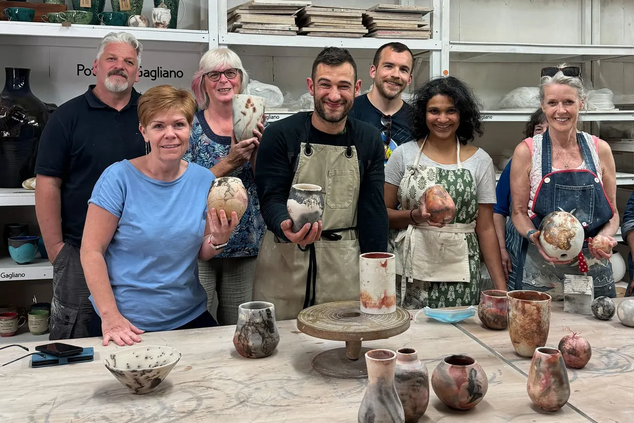 Group of seven people holding handmade pottery pieces in a studio workshop with shelves of ceramics in the background.