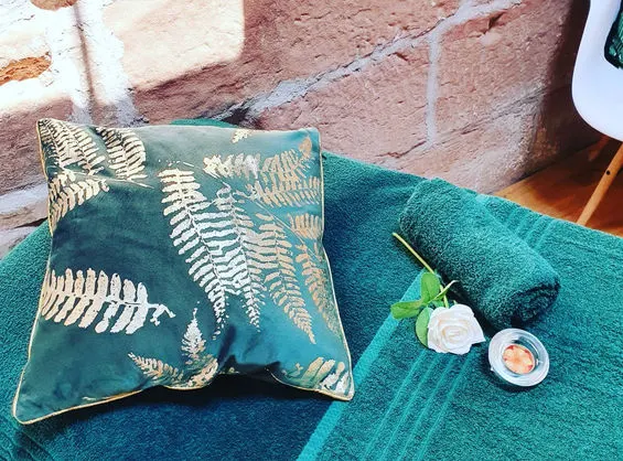 Teal cushion with gold fern design and small spa products arranged on matching towel in outdoor setting.