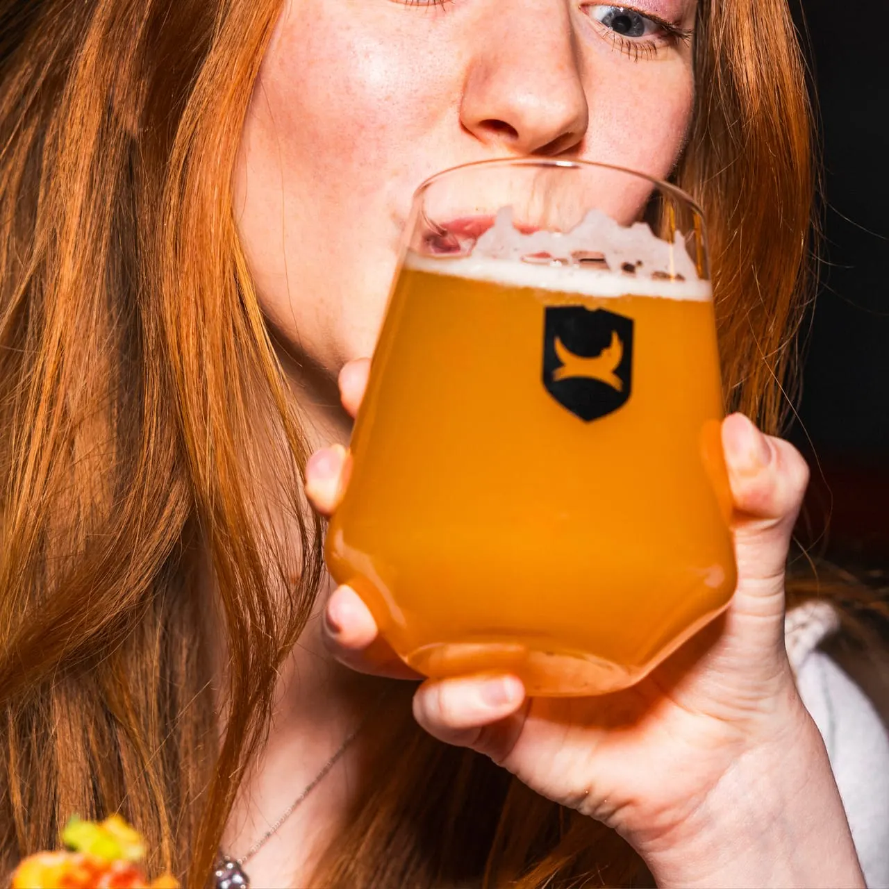 Woman with long reddish-brown hair drinking from a glass of golden ale with a branded logo.
