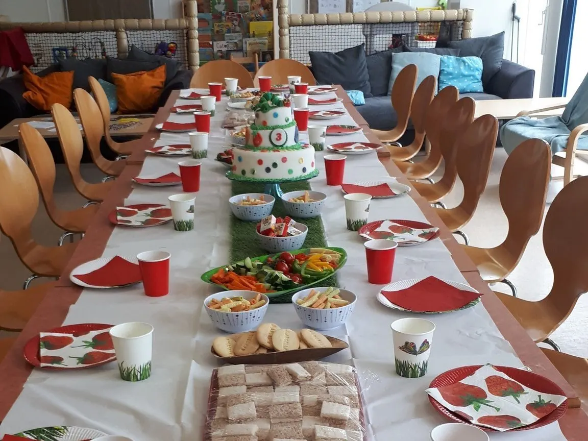 Long table set for a children's party with decorated cake, food, red cups and napkins, wooden chairs either side.