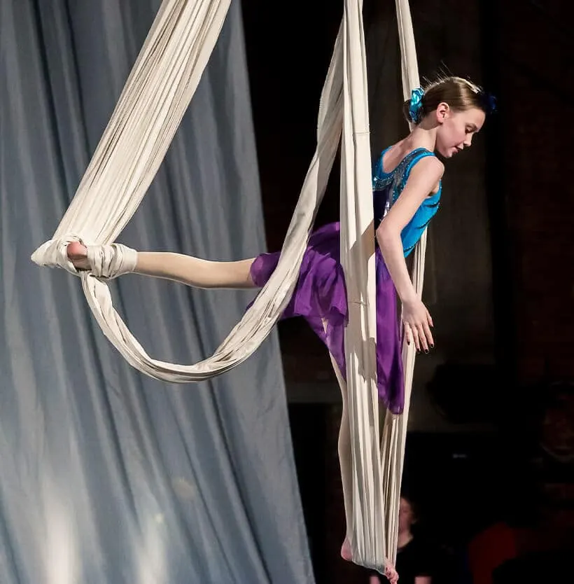 Young acrobat performing aerial silk routine suspended in white fabric against dark stage backdrop.
