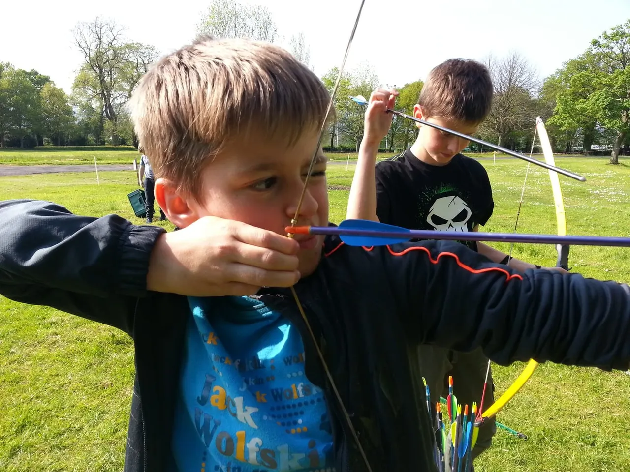 Two boys practising archery outdoors, aiming with bows on a grassy field with targets visible in the background.