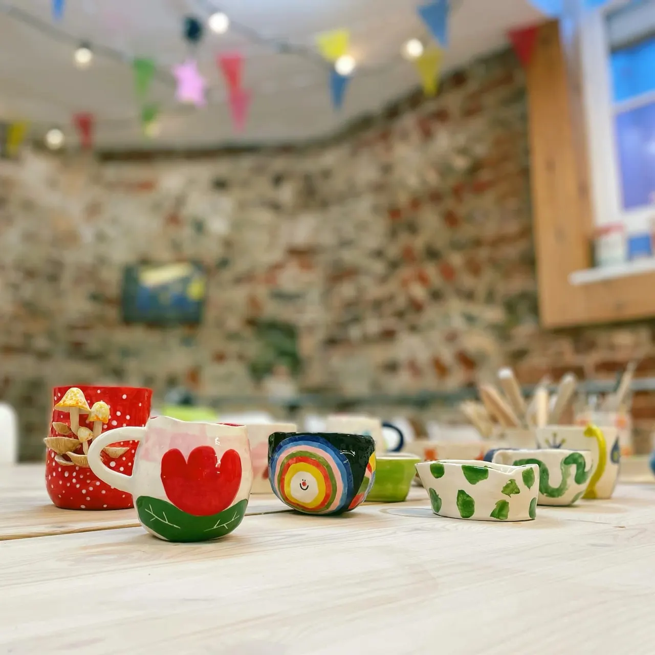 Colourful hand-painted ceramic bowls and mugs displayed on a wooden table in a bright craft studio with bunting and a stone feature wall.