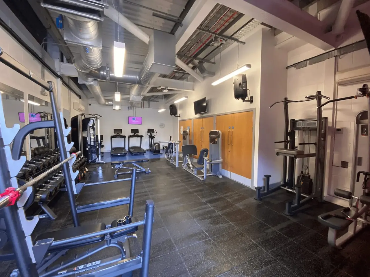 This is a photograph, not a designed graphic.

Modern gym interior with various exercise equipment including treadmills, weight machines, and dumbbells on dark flooring.