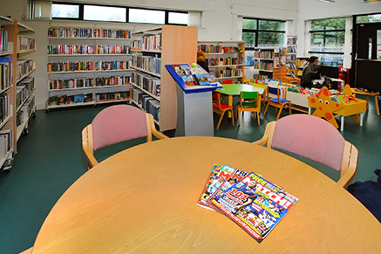 Children's library area with round wooden table, colourful chairs, bookshelves, and play equipment, with children visible in background.
