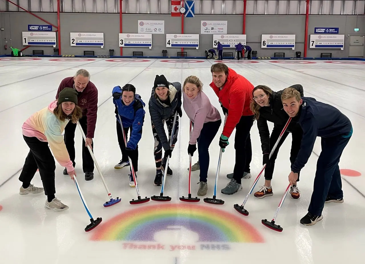 Group of eight people holding curling brushes and stones on an ice rink, posing around a rainbow design.
