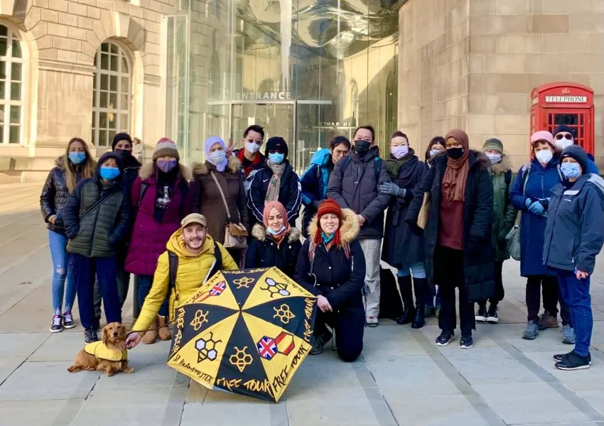 Large group of tourists wearing masks posing outside a building entrance with a red telephone box, holding a nautical-themed umbrella.