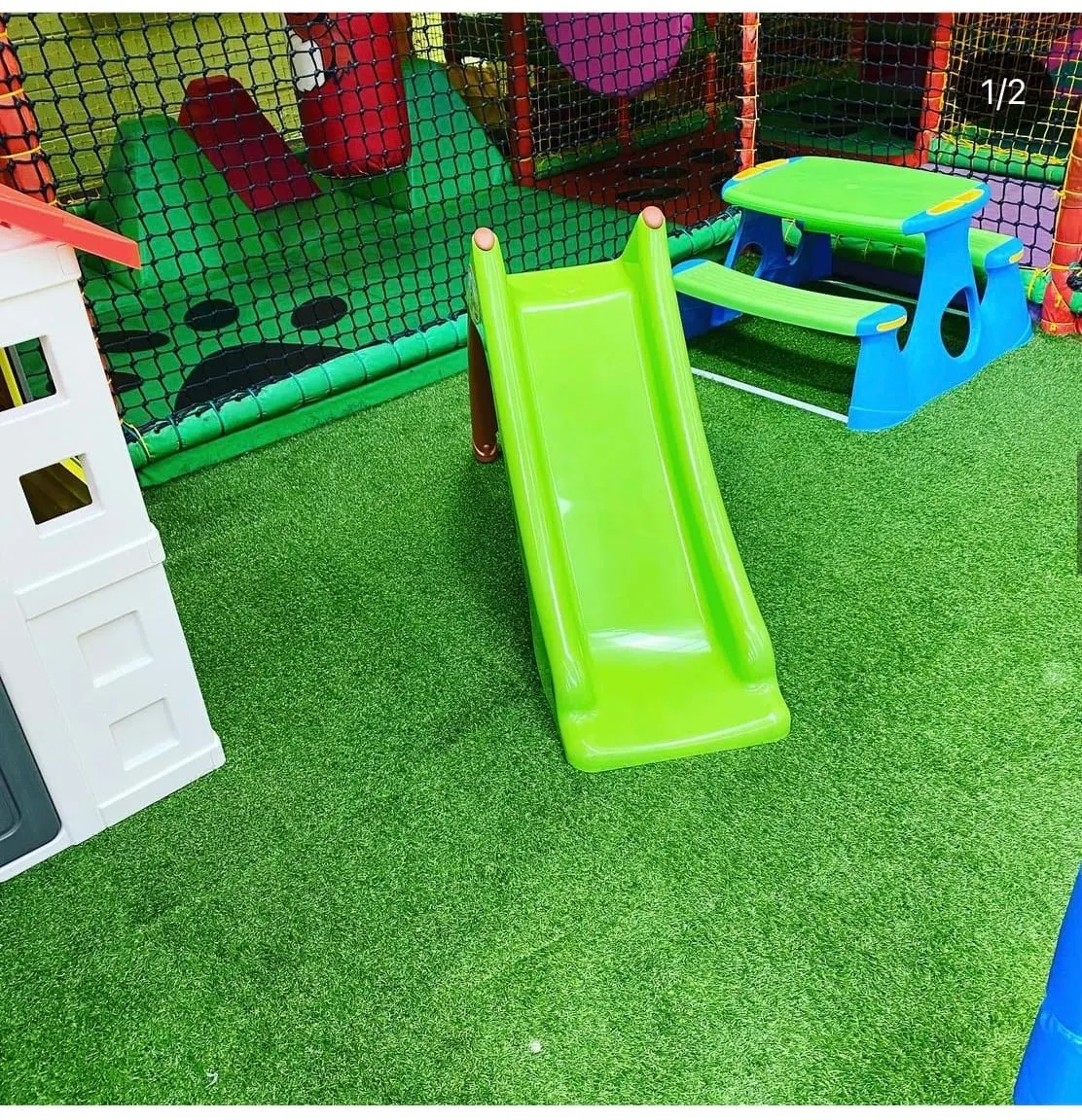 Bright lime-green slide and blue picnic table in an indoor soft play area with artificial turf and colourful netting.