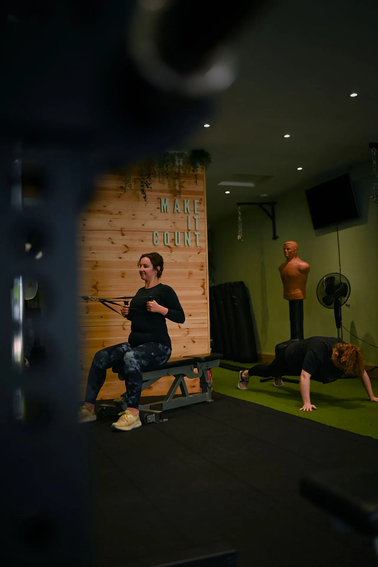 Two people exercising in a gym studio with green walls and wooden "Make It Count" signage.