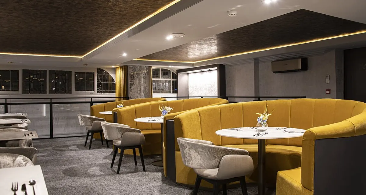 Modern restaurant interior with curved mustard-yellow banquette seating, small round tables, and contemporary lighting throughout the space.