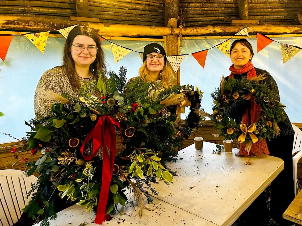 Three young people display festive wreaths decorated with greenery, red ribbons and dried fruit in a rustic barn setting.