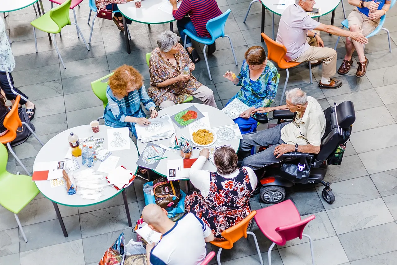 Overhead view of older adults seated at colourful chairs around tables, engaged in an art and craft activity together indoors.