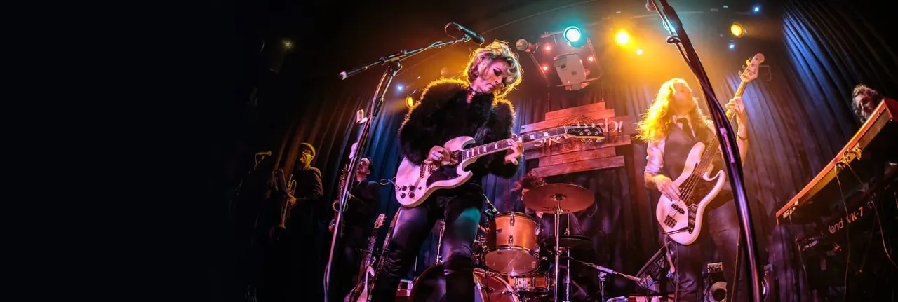 Rock band performing on stage with guitarist, bassist, drummer and other musicians under coloured stage lighting.