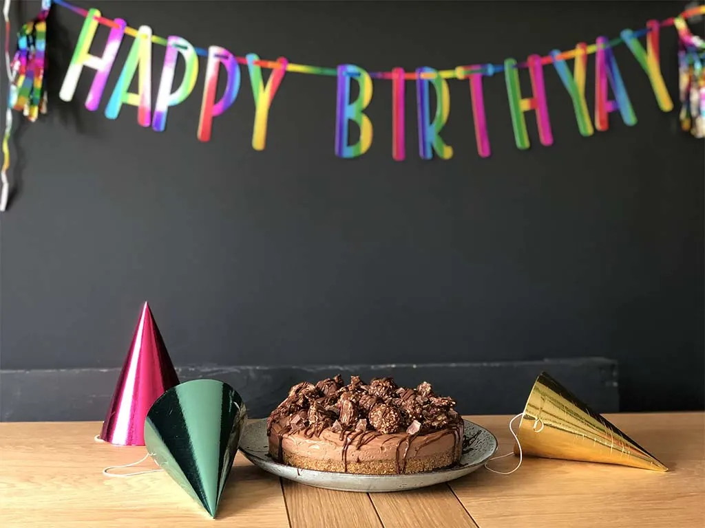 Colourful "Happy Birthday" bunting hangs above a wooden table decorated with party hats and a chocolate cake.